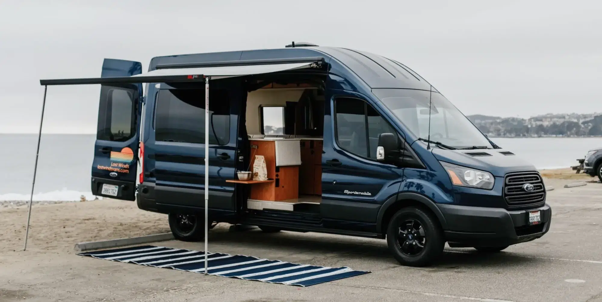 Dark blue Ford Transit with open doors parked next to a lake.