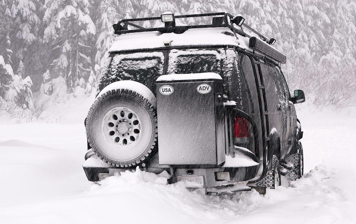 Black converted van driving through a forest on a snowy day.